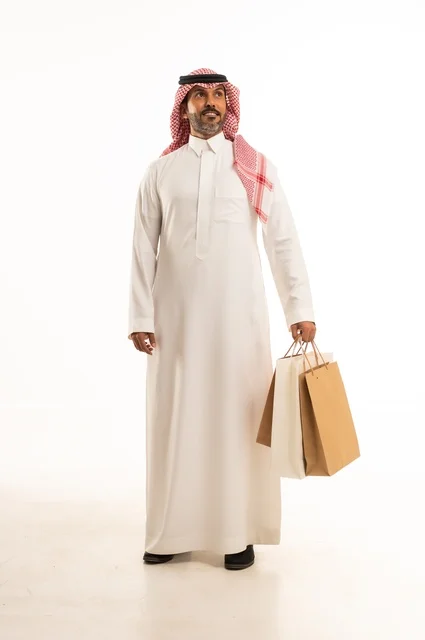 المتعة في التسوق واقتناء المستلزمات والهدايا، الاستفادة من العروضات والتخفيضات، بورتريه لرجل عربي خليجي سعودي يرتدي الشماغ والثوب التقليدي يحمل بيده أكياس ورقية ملونة بإيماءات السرور والسعادة، الجسم بالطول الكامل، خلفية بيضاء