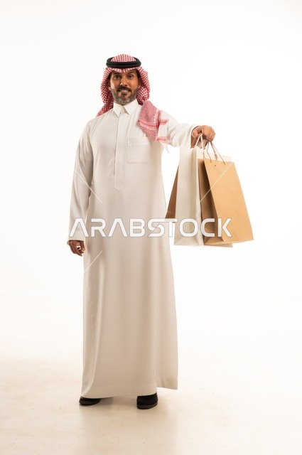 المتعة في التسوق واقتناء المستلزمات والهدايا، الاستفادة من العروضات والتخفيضات، بورتريه لرجل عربي خليجي سعودي يرتدي الشماغ والثوب التقليدي يحمل بيده أكياس ورقية ملونة بإيماءات السرور والسعادة، الجسم بالطول الكامل، خلفية بيضاء