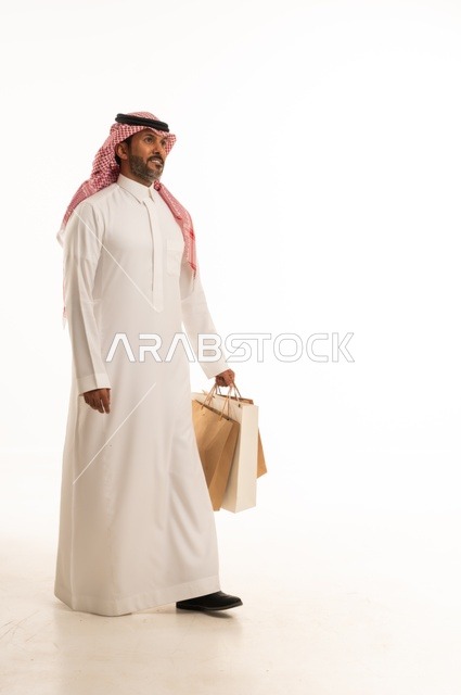 المتعة في التسوق واقتناء المستلزمات والهدايا، الاستفادة من العروضات والتخفيضات، بورتريه لرجل عربي خليجي سعودي يرتدي الشماغ والثوب التقليدي يحمل بيده أكياس ورقية ملونة بإيماءات السرور والسعادة، الجسم بالطول الكامل، خلفية بيضاء