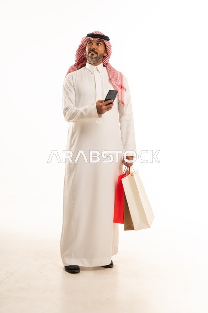Completing the ordering process using a mobile phone, the concept of remote shopping, online electronic purchases, a portrait of a young Arab Gulf Saudi man wearing traditional attire holding colorful paper bags, shopping and taking advantage of offers and discounts, full-length body, white background.