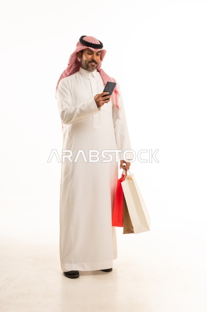 Completing the ordering process using a mobile phone, the concept of remote shopping, online electronic purchases, a portrait of a young Arab Gulf Saudi man wearing traditional attire holding colorful paper bags, shopping and taking advantage of offers and discounts, full-length body, white background.