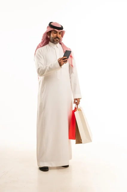Completing the ordering process using a mobile phone, the concept of remote shopping, online electronic purchases, a portrait of a young Arab Gulf Saudi man wearing traditional attire holding colorful paper bags, shopping and taking advantage of offers and discounts, full-length body, white background.