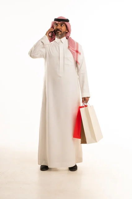 Making a call using a mobile phone, the concept of remote shopping, online electronic purchases, a portrait of a young Arab Gulf Saudi man wearing traditional attire holding colorful paper bags, shopping and benefiting from offers and discounts, full-length body, white background.