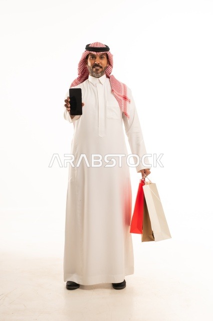 Displaying a blank black screen using a mobile phone, the concept of remote shopping, online electronic purchases, a portrait of a Saudi Arabian Gulf man wearing traditional attire holding colorful paper bags, shopping and taking advantage of offers and discounts, full-length body, white background.