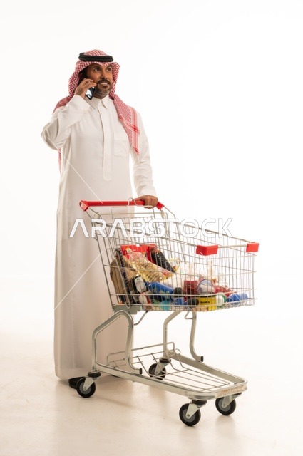 اجراء مكالمة هاتفية عبر الجوال، الاستمتاع بالتسوق في المراكز التجارية، شراء المستلزمات والحاجات الضرورية، الاستفادة من الخصومات على المنتجات، بورتريه لرجل عربي خليجي سعودي يرتدي الزي التقليدي ويجرعربة التسوق بإيماءات السعادة، الجسم بالطول الكامل، خلفية بيضاء