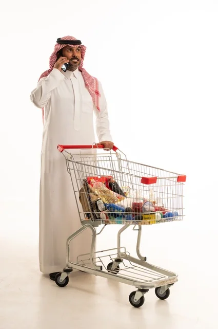 اجراء مكالمة هاتفية عبر الجوال، الاستمتاع بالتسوق في المراكز التجارية، شراء المستلزمات والحاجات الضرورية، الاستفادة من الخصومات على المنتجات، بورتريه لرجل عربي خليجي سعودي يرتدي الزي التقليدي ويجرعربة التسوق بإيماءات السعادة، الجسم بالطول الكامل، خلفية بيضاء