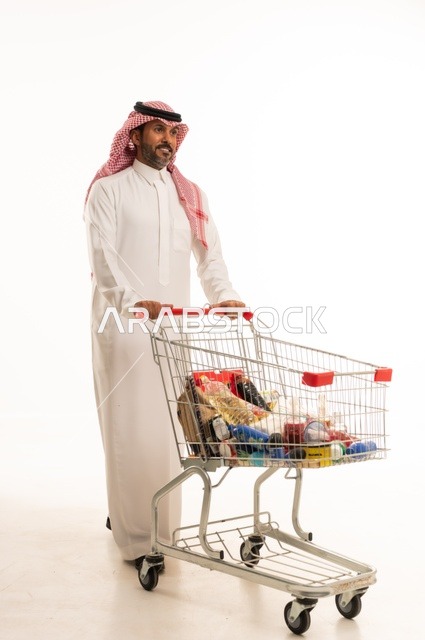 Enjoying shopping in malls and commercial centers, buying necessities and essential items, taking advantage of discounts on products, a portrait of a Saudi Arabian Gulf man wearing traditional attire and a ghutrah, pulling a shopping cart in front of him with gestures of happiness, full-body, white background.