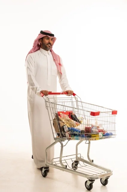 Enjoying shopping in malls and commercial centers, buying necessities and essential items, taking advantage of discounts on products, a portrait of a Saudi Arabian Gulf man wearing traditional attire and a ghutrah, pulling a shopping cart in front of him with gestures of happiness, full-body, white background.