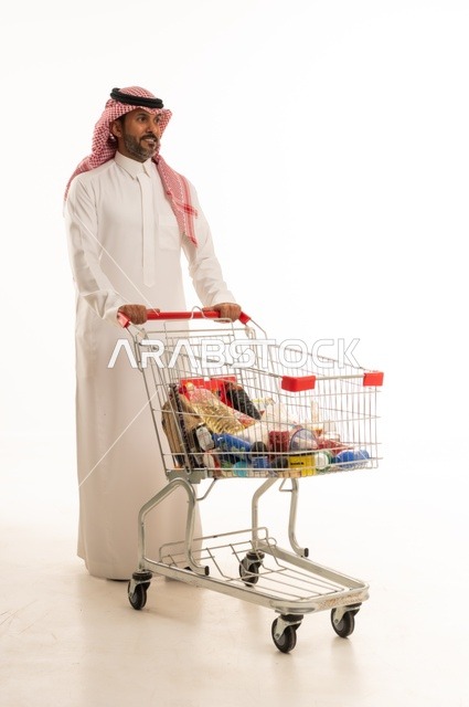 الاستمتاع بالتسوق في المولات والمراكز التجارية، شراء المستلزمات والحاجيات الضرورية، الاستفادة من الخصومات على المنتجات، بورتريه لرجل عربي خليجي سعودي يرتدي الثوب التقليدي و الشماغ ويجر عربة التسوق أمامه بإيماءات السعادة، الجسم بالطول الكامل، خلفية بيضاء
