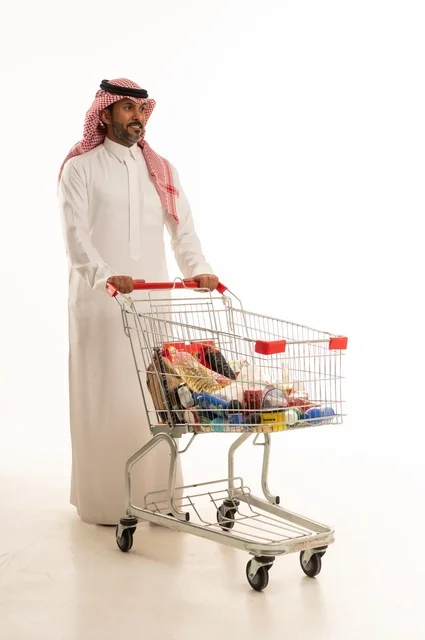 الاستمتاع بالتسوق في المولات والمراكز التجارية، شراء المستلزمات والحاجيات الضرورية، الاستفادة من الخصومات على المنتجات، بورتريه لرجل عربي خليجي سعودي يرتدي الثوب التقليدي و الشماغ ويجر عربة التسوق أمامه بإيماءات السعادة، الجسم بالطول الكامل، خلفية بيضاء