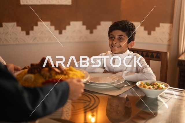 التعرف على أداء فريضة الصيام في شهر رمضان، طفل عربي خليجي سعودي يرتدي ثوب أبيض في موعد كسر الصيام عند الأذان، أجواء عائلية روحانية دينية إسلامية، ايماءات السرور لاجتماع العائلة حول مائدة إفطار رمضان، سفرة مليئة بأصناف من المأكولات الشرقية الشهية