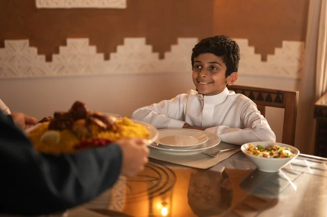 Recognizing the performance of the fasting obligation during the month of Ramadan, a Saudi Arabian Gulf Arab child wearing a white thobe at the time of breaking the fast at the call to prayer, a spiritual and religious Islamic family atmosphere, gestures of joy for the family gathering around the Ramadan iftar table, a spread full of delicious Eastern dishes.