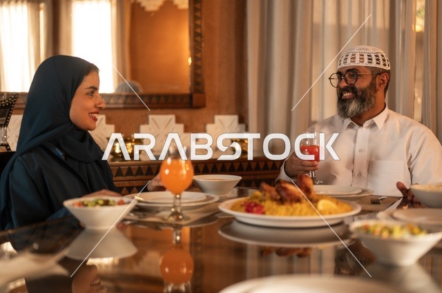Performing the obligation of fasting in the blessed month of Ramadan, happy religious family Ramadan atmosphere, exchanging conversations among family members, an Arab Gulf Saudi family gathered around the dining table, a spread full of delicious Eastern dishes, decorations for Islamic occasions.