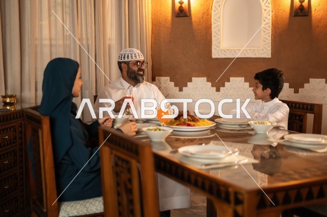 أجواء عائلية دينية رمضانية سعيدة، تبادل الأحاديث بين أفراد الأسرة، عائلة عربية خليجية سعودية مجتمعة حول مائدة الطعام، سفرة مليئة بأصناف من المأكولات الشرقية الشهية، أداء فريضة الصيام في شهر رمضان المبارك، زينة المناسبات الإسلامية