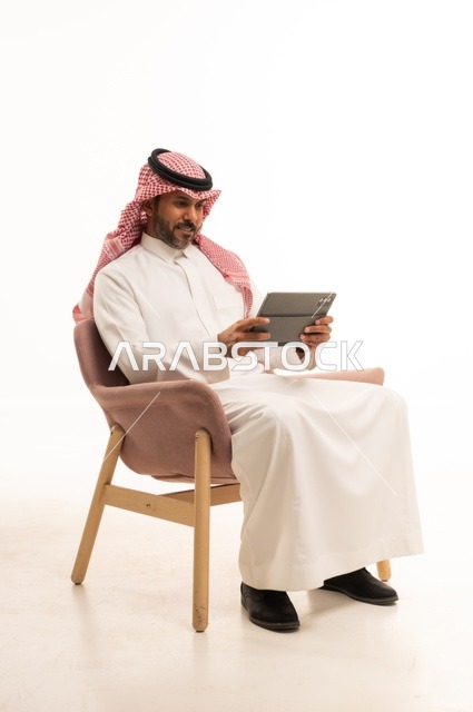 Chatting with family and friends, using modern technology and techniques, a portrait of an Arab Gulf Omani man wearing traditional thobe and shmagh holding a tablet in his hand, browsing social media, using applications and programs on tablet devices, white background.