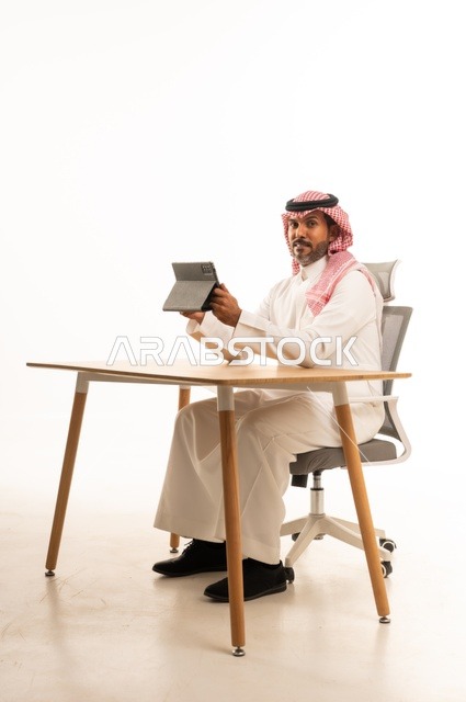 انجاز مهام العمل عبر الانترنت، استعمال التكنولوجيا والتقنيات الحديثة، بورتريه لرجل عربي خليجي عماني يرتدي الثوب والشماغ التقليدي يجلس خلف الطاولة يحمل بيده جهاز التابلت، تصفح مواقع التواصل الاجتماعي، استخدام تطبيقات وبرامج الأجهزة اللوحية، خلفية بيضاء