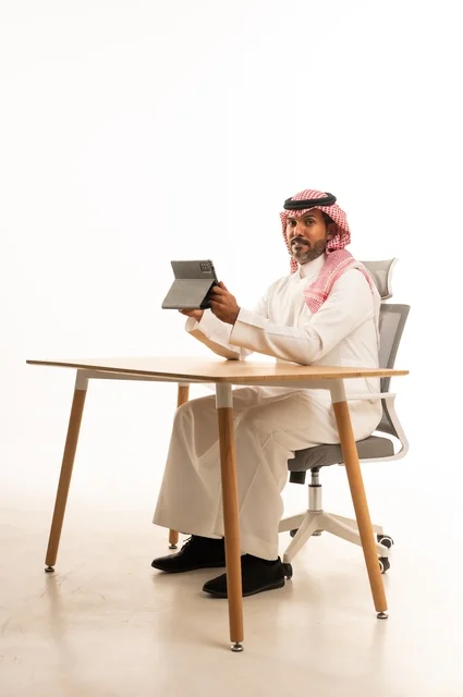 انجاز مهام العمل عبر الانترنت، استعمال التكنولوجيا والتقنيات الحديثة، بورتريه لرجل عربي خليجي عماني يرتدي الثوب والشماغ التقليدي يجلس خلف الطاولة يحمل بيده جهاز التابلت، تصفح مواقع التواصل الاجتماعي، استخدام تطبيقات وبرامج الأجهزة اللوحية، خلفية بيضاء