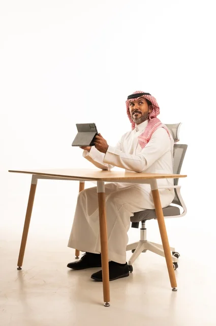 انجاز مهام العمل عبر الانترنت، استعمال التكنولوجيا والتقنيات الحديثة، بورتريه لرجل عربي خليجي عماني يرتدي الثوب والشماغ التقليدي يجلس خلف الطاولة يحمل بيده جهاز التابلت، تصفح مواقع التواصل الاجتماعي، استخدام تطبيقات وبرامج الأجهزة اللوحية، خلفية بيضاء