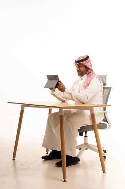 انجاز مهام العمل عبر الانترنت، استعمال التكنولوجيا والتقنيات الحديثة، بورتريه لرجل عربي خليجي عماني يرتدي الثوب والشماغ التقليدي يجلس خلف الطاولة يحمل بيده جهاز التابلت، تصفح مواقع التواصل الاجتماعي، استخدام تطبيقات وبرامج الأجهزة اللوحية، خلفية بيضاء