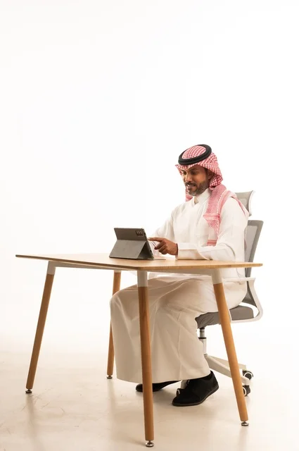 Completing work tasks online, using technology and modern techniques, a portrait of an Arab Gulf Omani man wearing traditional thobe and shmagh sitting behind a table holding a tablet, browsing social media, using applications and programs for tablets, white background.
