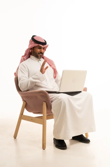 Completing work tasks online, using modern technology and techniques, a portrait of an Arab Gulf Omani man wearing traditional thobe and shmagh sitting on a comfortable chair holding a laptop, browsing social media, using mobile applications and programs, white background.