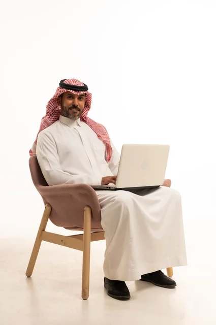 Completing work tasks online, using modern technology and techniques, a portrait of an Arab Gulf Omani man wearing traditional thobe and shmagh sitting on a comfortable chair holding a laptop, browsing social media, using mobile applications and programs, white background.