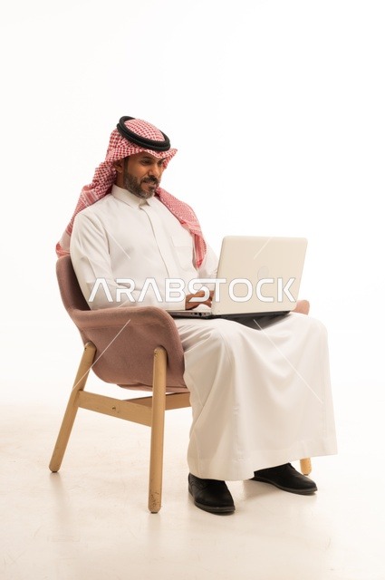 Completing work tasks online, using modern technology and techniques, a portrait of an Arab Gulf Omani man wearing traditional thobe and shmagh sitting on a comfortable chair holding a laptop, browsing social media, using mobile applications and programs, white background.