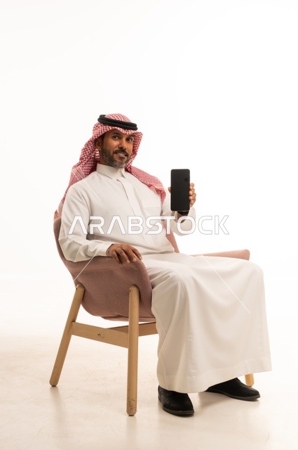 عرض شاشة سوداء فارغة عبر الهاتف المحمول، استعمال التكنولوجيا والتقنيات الحديثة، بورتريه لرجل عربي خليجي عماني يرتدي الزي التقليدي يجلس على كرسي مريح يحمل بيده جهاز الجوال، تصفح مواقع التواصل الاجتماعي، استخدام تطبيقات الأجهزة المحمولة، خلفية بيضاء
