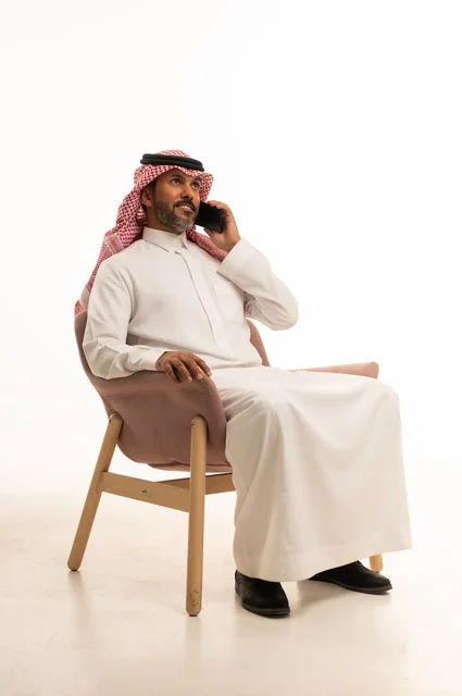 Making a phone call using a mobile phone, using modern technology and techniques, a portrait of an Arab Gulf Omani man wearing traditional attire sitting on a comfortable chair holding a mobile device, browsing social media, using mobile applications, white background.