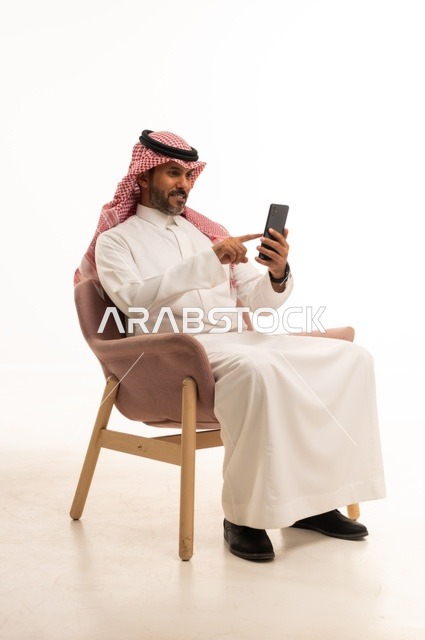 استعمال التكنولوجيا والتقنيات الحديثة، بورتريه لرجل عربي خليجي عماني يرتدي الزي التقليدي يجلس على كرسي مريح يحمل بيده جهاز الجوال، تصفح مواقع التواصل الاجتماعي، استخدام تطبيقات الأجهزة المحمولة، خلفية بيضاء