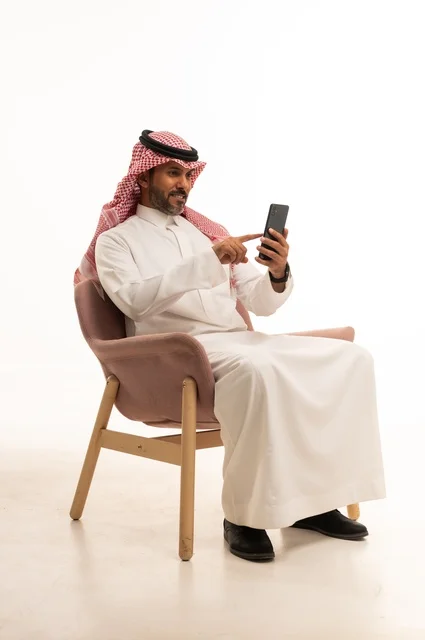 The use of technology and modern techniques, a portrait of an Arab Gulf Omani man wearing traditional attire sitting on a comfortable chair holding a mobile device, browsing social media, using mobile applications, white background.