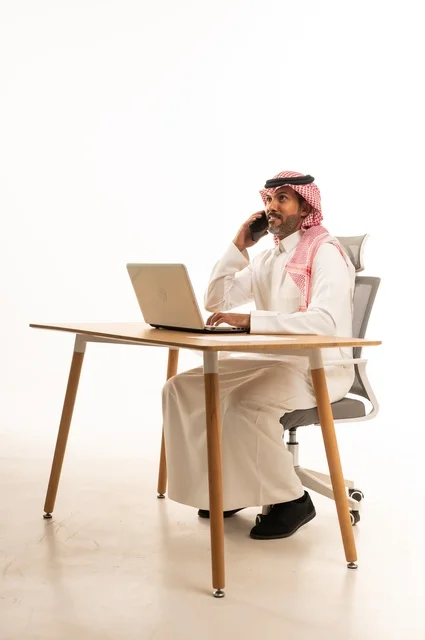 التواصل مع العملاء عبر الهاتف، انجاز مهام العمل عبر الانترنت، استعمال التكنولوجيا والتقنيات الحديثة، بورتريه لرجل عربي خليجي عماني يرتدي الزي التقليدي يجلس خلف يحمل بيده جهاز اللابتوب، تصفح مواقع التواصل الاجتماعي، استخدام تطبيقات الأجهزة المحمولة، خلفية بيضاء