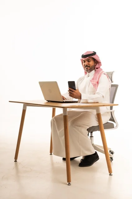التواصل مع العملاء عبر الهاتف، انجاز مهام العمل عبر الانترنت، استعمال التكنولوجيا والتقنيات الحديثة، بورتريه لرجل عربي خليجي عماني يرتدي الزي التقليدي يجلس خلف يحمل بيده جهاز اللابتوب، تصفح مواقع التواصل الاجتماعي، استخدام تطبيقات الأجهزة المحمولة، خلفية بيضاء