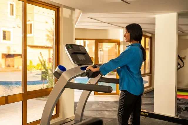 Using sports equipment and doing warm-up exercises, an Arab Gulf Saudi woman wearing sportswear is exercising in the gym, engaging in sports activities and exercises, health and fitness, with a background of a natural scene featuring green plants and a water pond.