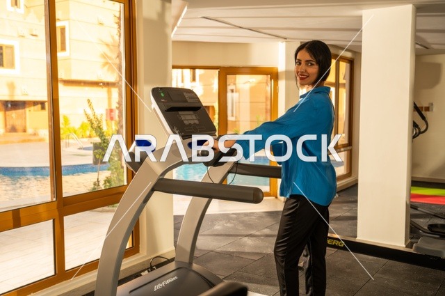 استخدام الأجهزة الرياضية والقيام بتمارين الإحماء ،  امرأة عربية خليجية سعودية ترتدي الملابس الرياضية وتقوم بالتمارين الرياضية في صالة الألعاب الرياضية، انشطة وتمارين رياضية ، الصحة واللياقة البدنية، خلفية منظر طبيعي نباتات خضراء مع بركة ماء