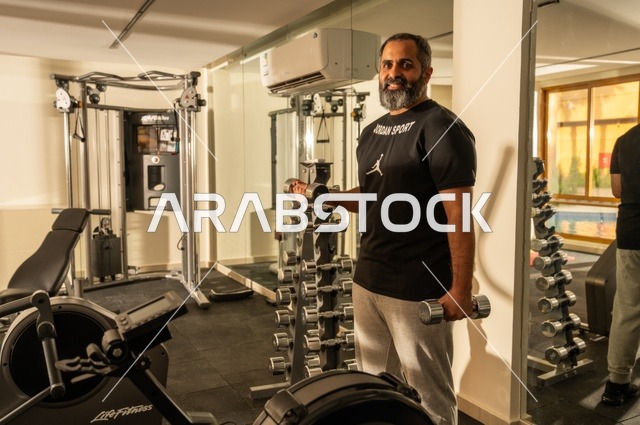 Gestures of strength and determination, attention to health and physical fitness, bodybuilding, using weights, an Arab Gulf Saudi man wearing sportswear practicing weightlifting exercises, a Saudi man holding weights in his hand, building muscle mass in the body.