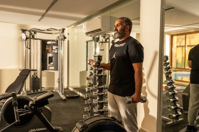 بناء الكتلة العضلية للجسم، إيماءات القوة والعزم، الاهتمام بالصحة واللياقة الجسدية، رياضة كمال الأجسام، استخدام الاثقال الرياضية، رجل عربي خليجي سعودي يرتدي الزي الرياضي يقوم بممارسة تمارين رفع الأوزان، رجل سعودي يحمل بيده الأوزان