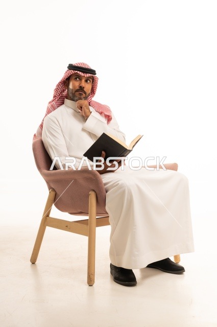 Spending time reading, learning, and culture, gestures of contemplation and focus, the concept of self-learning, a portrait of an Arab Saudi Gulf man wearing traditional Saudi attire sitting on a comfortable chair reading a book, white background.