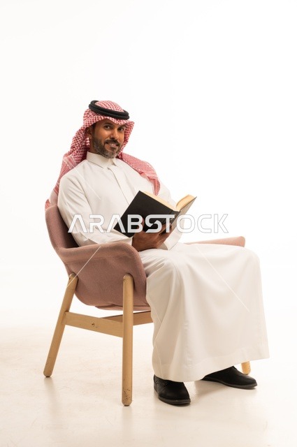 Spending time reading, learning, and culture, gestures of contemplation and focus, the concept of self-learning, a portrait of an Arab Saudi Gulf man wearing traditional Saudi attire sitting on a comfortable chair reading a book, white background.