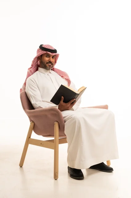 Spending time reading, learning, and culture, gestures of contemplation and focus, the concept of self-learning, a portrait of an Arab Saudi Gulf man wearing traditional Saudi attire sitting on a comfortable chair reading a book, white background.