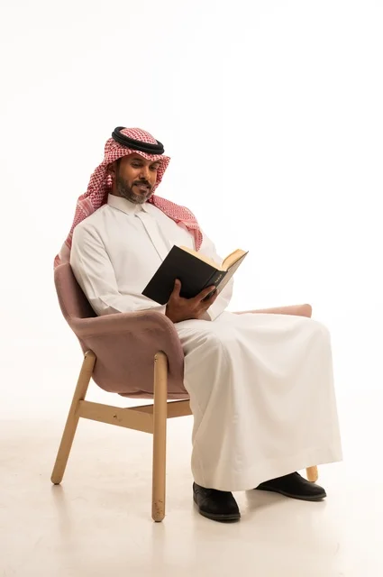 Spending time reading, learning, and culture, gestures of contemplation and focus, the concept of self-learning, a portrait of an Arab Saudi Gulf man wearing traditional Saudi attire sitting on a comfortable chair reading a book, white background.