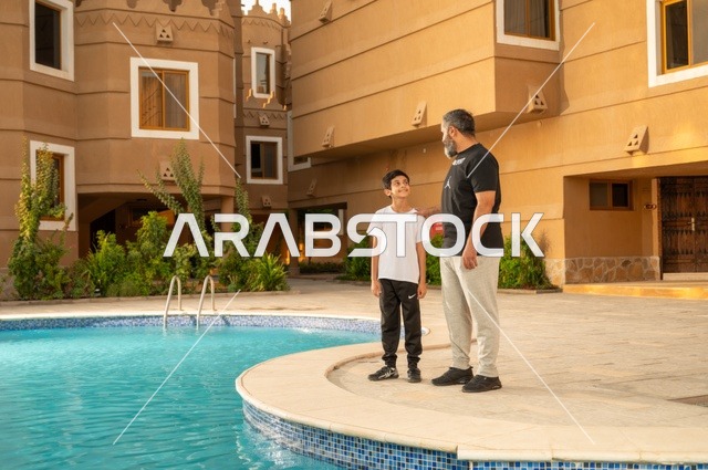 مشاركة العائلة اللحظات الممتعة, القيام بالأنشطة الترفيهية المسلية, قضاء العطلة الصيفية في المنتجعات السياحية, اب عربي خليجي سعودي يقف بجانب حوض السباحة مع ولده في إحدى الفنادق بالسعودي، ممارسة رياضة السباحة الممتعة