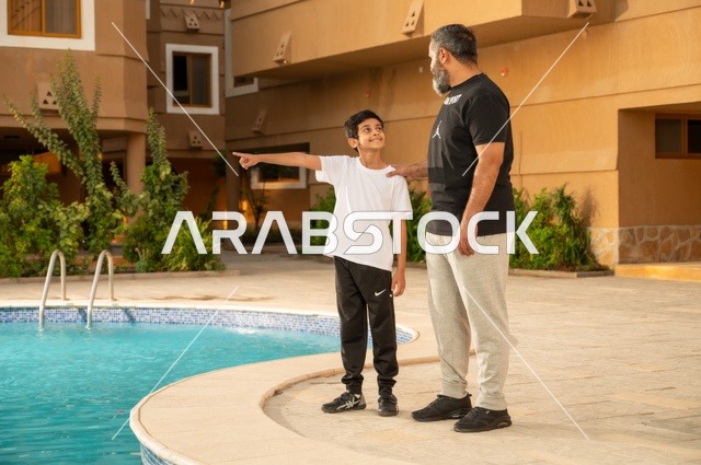 الإشارة إلى شيء ما، قضاء العطلة الصيفية في المنتجعات السياحية, مشاركة العائلة اللحظات الممتعة, القيام بالأنشطة الترفيهية المسلية, اب عربي خليجي سعودي يقف بجانب حوض السباحة مع ولده في إحدى الفنادق بالسعودي، ممارسة رياضة السباحة الممتعة