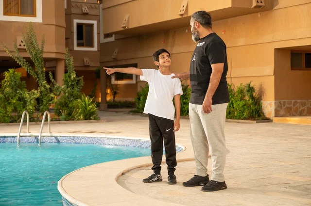 الإشارة إلى شيء ما، قضاء العطلة الصيفية في المنتجعات السياحية, مشاركة العائلة اللحظات الممتعة, القيام بالأنشطة الترفيهية المسلية, اب عربي خليجي سعودي يقف بجانب حوض السباحة مع ولده في إحدى الفنادق بالسعودي، ممارسة رياضة السباحة الممتعة