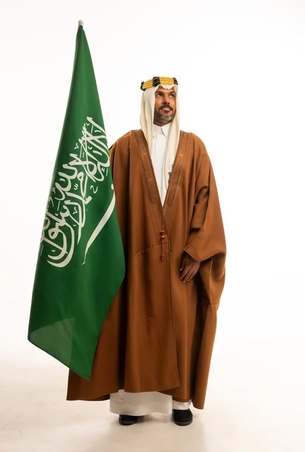 الفخر والاعتزاز بالهوية الوطنية، اليوم الوطني السعودي 23 سبتمبر، بورتريه لرجل عربي خليجي سعودي مبتسم يرتدي الالبشت وعقال التأسيس يحمل بيده علم المملكة، الاحتفال بيوم العلم 11 مارس، صورة الجسم بالكامل، خلفية بيضاء