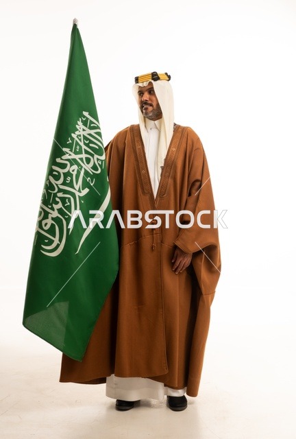 الفخر والاعتزاز بالهوية الوطنية، اليوم الوطني السعودي 23 سبتمبر، بورتريه لرجل عربي خليجي سعودي مبتسم يرتدي الالبشت وعقال التأسيس يحمل بيده علم المملكة، الاحتفال بيوم العلم 11 مارس، صورة الجسم بالكامل، خلفية بيضاء