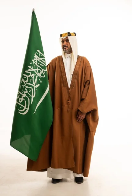 Pride and honor in national identity, Saudi National Day September 23, a portrait of a smiling Saudi Gulf Arab man wearing a bisht and the founding agal holding the Kingdom's flag in his hand, celebrating Flag Day March 11, full-body image, white background.