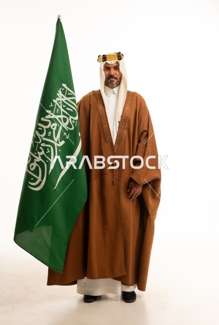 الفخر والاعتزاز بالهوية الوطنية، اليوم الوطني السعودي 23 سبتمبر، بورتريه لرجل عربي خليجي سعودي مبتسم يرتدي الالبشت وعقال التأسيس يحمل بيده علم المملكة، الاحتفال بيوم العلم 11 مارس، صورة الجسم بالكامل، خلفية بيضاء