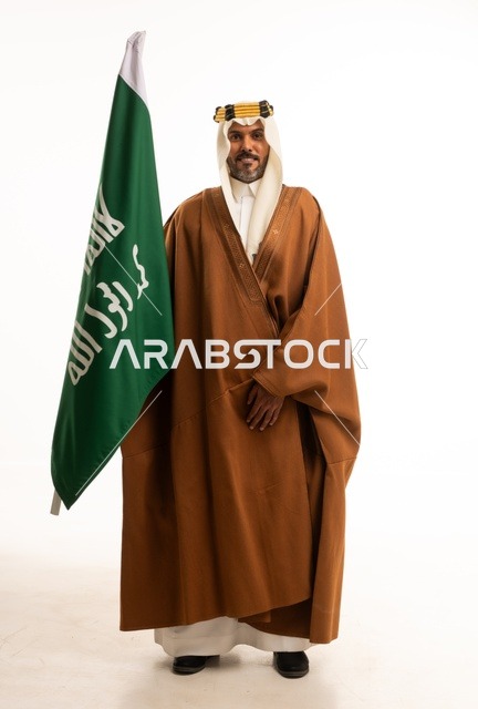 اليوم الوطني السعودي 23 سبتمبر، الفخر والاعتزاز بالهوية الوطنية، بورتريه لرجل عربي خليجي سعودي مبتسم يرتدي البشت وعقال التأسيس يحمل بيده علم تأسيس المملكة السعودية 22 فبراير، الاحتفال بيوم بدينا 1727 ، صورة الجسم بالكامل، خلفية بيضاء