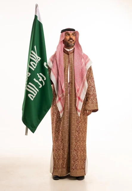 Wearing traditional attire on national occasions, Saudi National Day on September 23, pride and honor for the homeland, a portrait of a Saudi Arabian Gulf man wearing a ghutrah and thobe holding in his hand the flag of the establishment of the Kingdom of Saudi Arabia on February 22, celebrating the Day of Bidaya 1727, full-body image, white background.